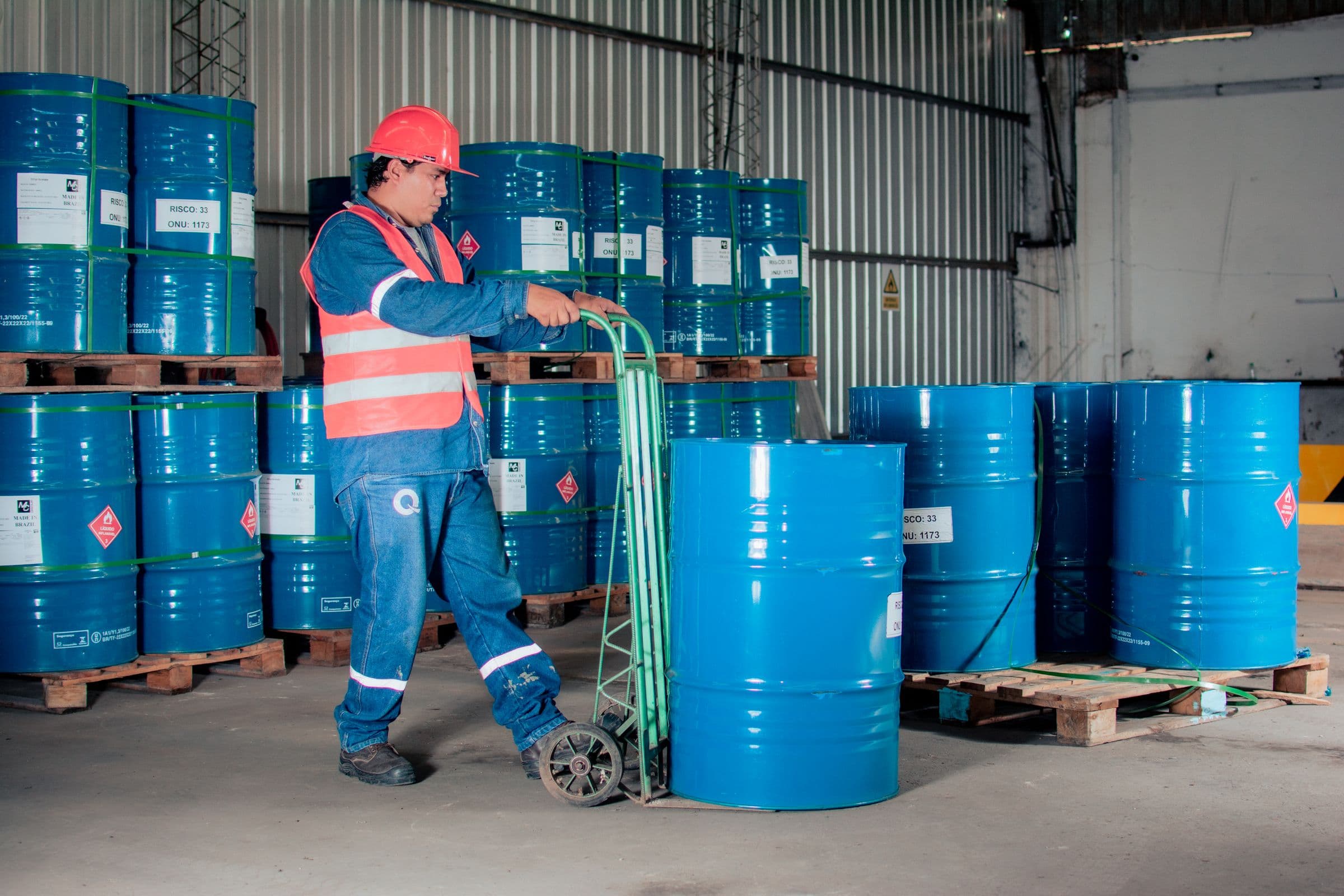 A worker in PPE moving blue dangerous goods drums, marked with UN hazard labels, in an industrial warehouse