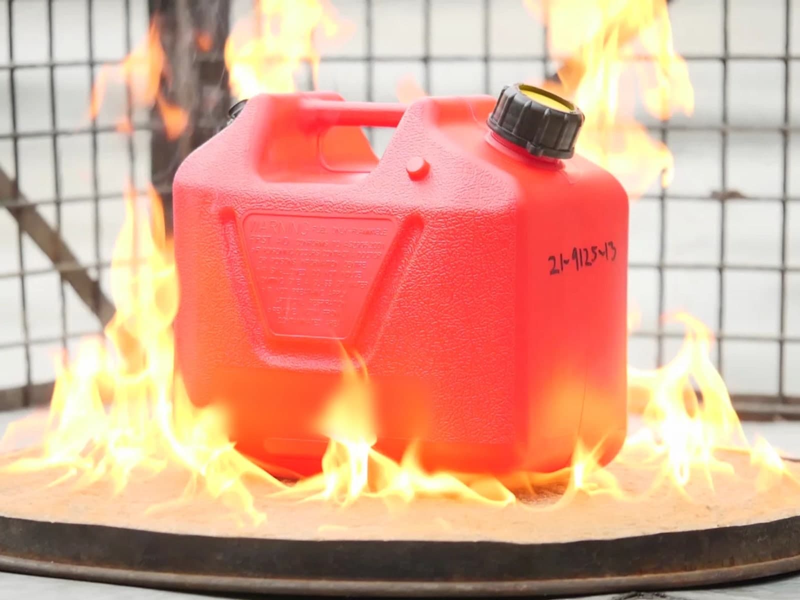 A red portable fuel container engulfed in flames on a steel test pad inside a wire-mesh cage during a fire test